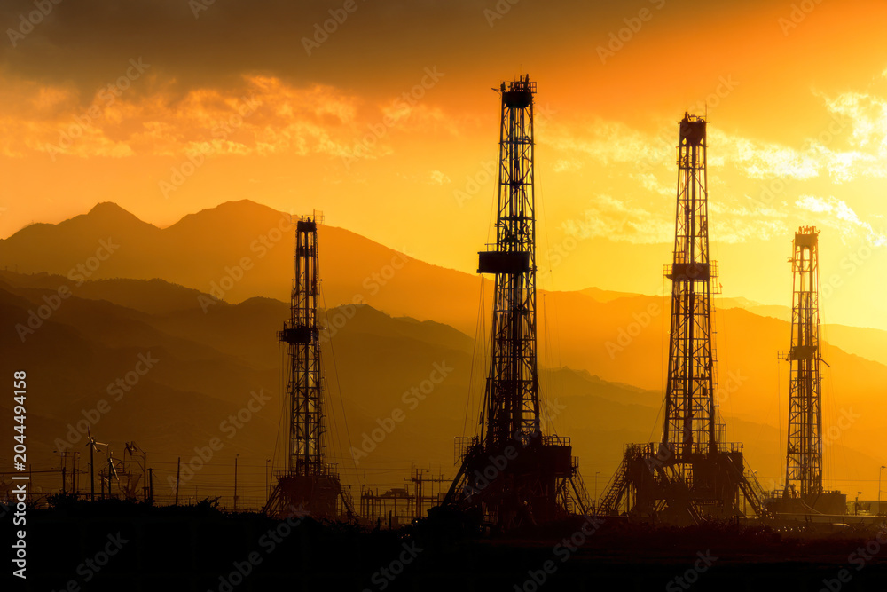 Obraz premium Silhouettes of oil and natural gas drilling rigs stand in front of mountains as the sun sets, creating a colorful sky. The scene shows energy production in a natural setting