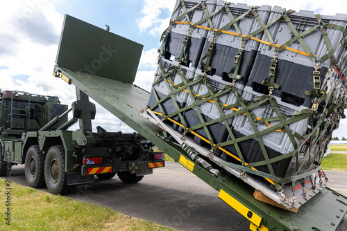 Military hook-lift truck unloads cargo pallet secured netting loading ramp airfield. Airforce logistics operation transport, deployment readiness, mobility support and defense infrastructure