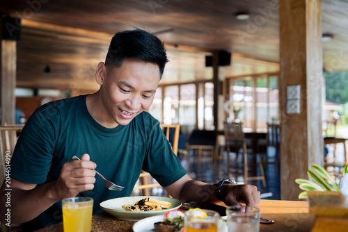 Asian man sits alone at a cozy wooden cafe, enjoying pancakes and orange juice for a relaxed breakfast.