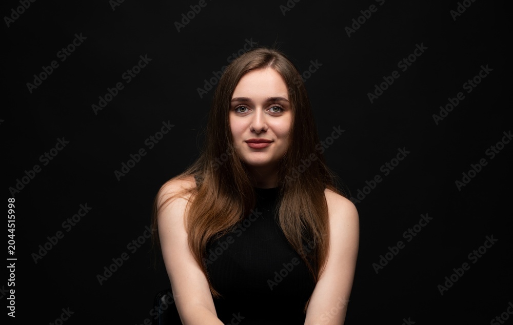 Naklejka premium Young woman with long brown hair smiling at camera, sitting on a chair against a black background