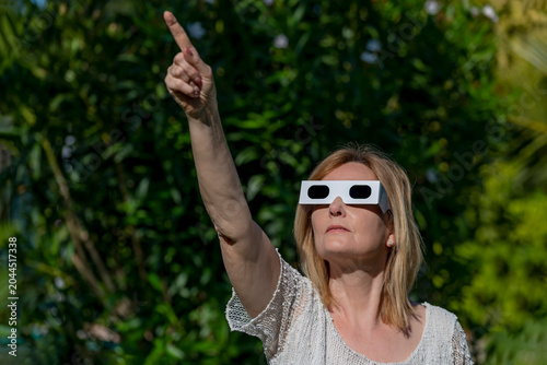 A woman wears glasses to observe a solar eclipse and points her index finger at the sun