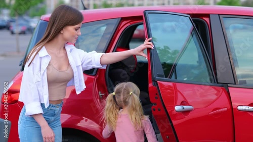 Smiling woman holding hand of little girl while walking on the parking to the car. Kid sitting in the car seat in the back seat. Family, transport, trip, safety and parenthood concept. Real time