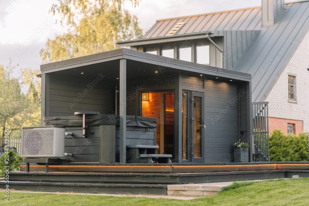 Obraz premium Modern wooden sauna with hot tub and outdoor deck situated in a residential backyard surrounded by greenery and a contemporary house in the background
