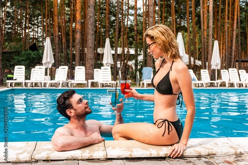 Fotografía Blonde woman in the bikini seated on the swimming pool edge clinking glasses wit