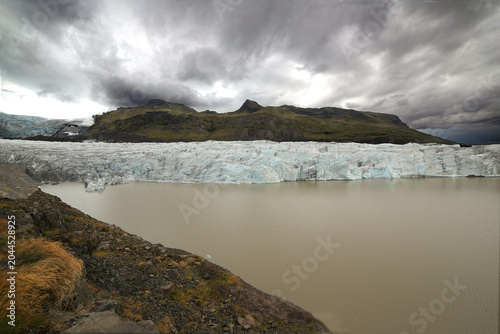 Fotografía In a national park in Iceland, a brownish glacial lake fed by melting ice is seen, contrasting with the blue ice, black volcanic ash and mountainous landscape