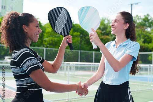 Two diverse young women friends, black and caucasian, in athletic wear smiling, holding pickleball paddle up and shaking hands over the net after match on modern outdoor court