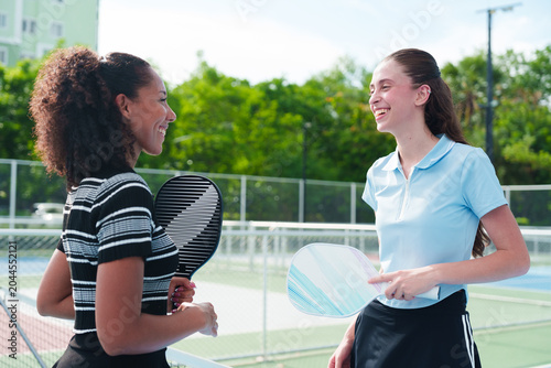 Two diverse young women friends, black and caucasian, smiling, holding pickleball paddle and talking over the net after match on modern outdoor court