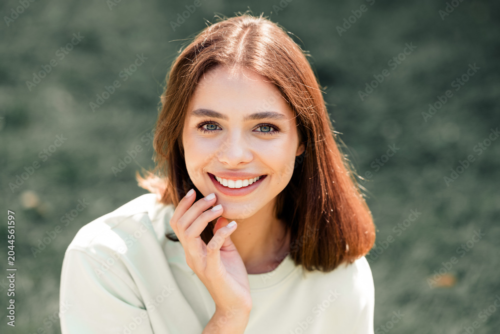 Fototapeta premium Smiling young woman portrait outdoors looking at camera with hand near face, natural light and casual clothing conveying friendly, confident lifestyle and wellness mood