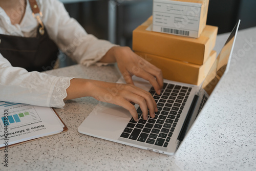 Hands typing on laptop keyboard managing online store inventory and shipping orders near parcel boxes and sales marketing report within office workspace