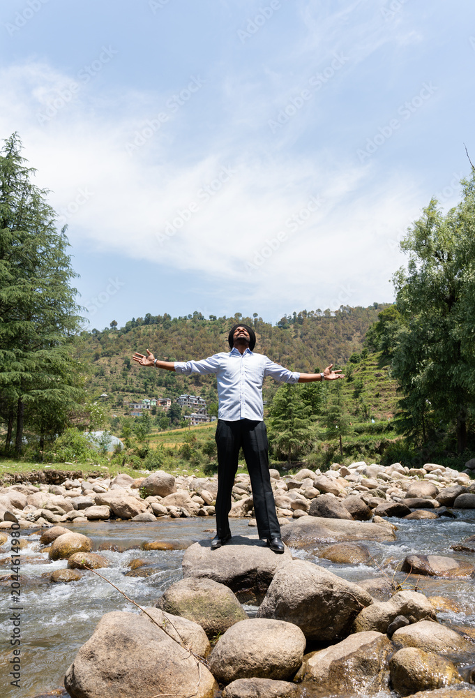 Obraz premium A vertical shot of a young Punjabi Sikh man wearing a black turban, standing with arms outstretched on a rock in a mountain river, looking up at the sky.