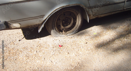 Foto Old rusty silver car with a completely flat rear tire, parked on a dusty, dry dirt road covered in debris