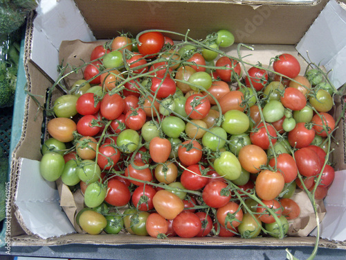 Top-down view of a cardboard box filled with fresh cherry tomatoes on the vine, showing a vibrant mix of ripe red, ripening orange, and unripe green colors
