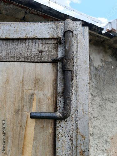 Close-up shot of a rustic, handmade metal slide latch locking a weathered wooden door inside a rusty iron frame. The old latch mechanism shows signs of aging, heavy use, and outdoor elements