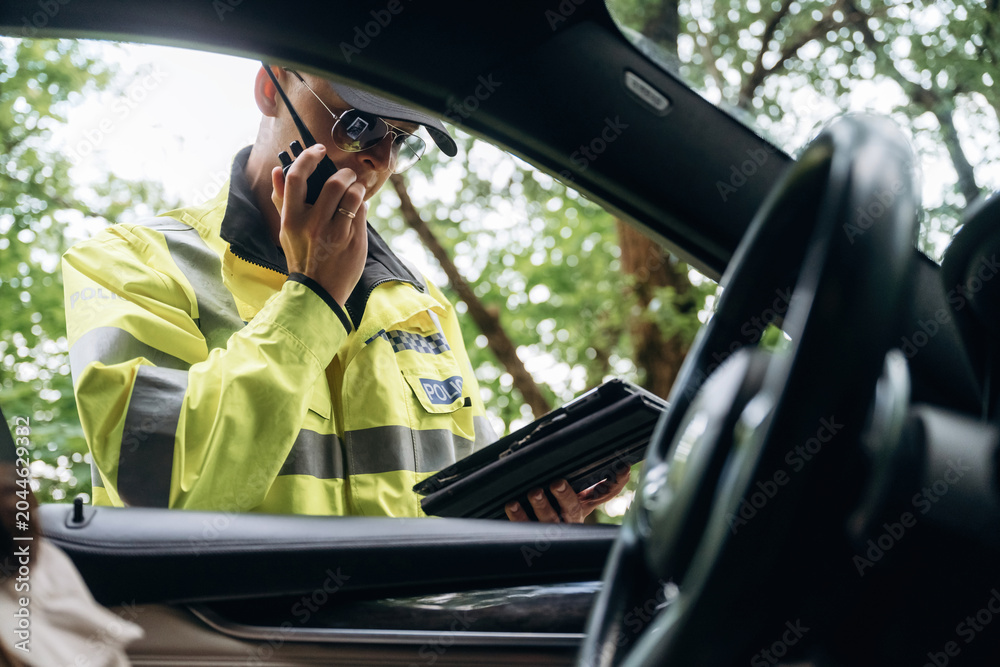 Obraz premium Male traffic officer in reflective yellow uniform using a smartphone and tablet while standing outside a car with blurred trees in the background