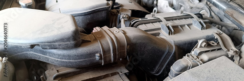 Detailed close-up shot of a dirty, dusty car engine bay, featuring a large black plastic air intake hose connected to a filter box. Various mechanical and electrical parts are visible