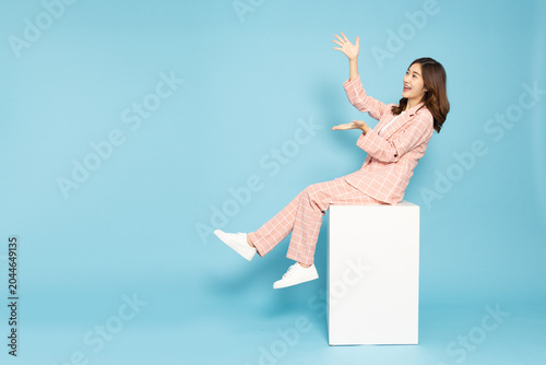 Happy Asian business woman sitting on a white cube, presenting or reaching out her hands. Cheerful female in a pink grid suit looking up at blank copy space, isolated on a blue studio background