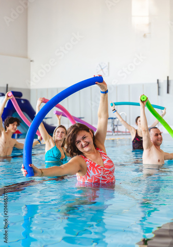 Group of people stretching with colorful noodles during an aqua aerobics class in an indoor swimming pool