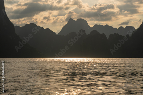 Bild auf Leinwand Golden sun glints dance on rippling water at sunset, silhouetting dramatic karst mountains against a cloudy sky in a serene natural landscape
