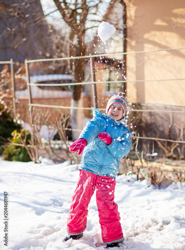 Smiling child colorful winter clothes standing on snow with trees and houses in sunny background