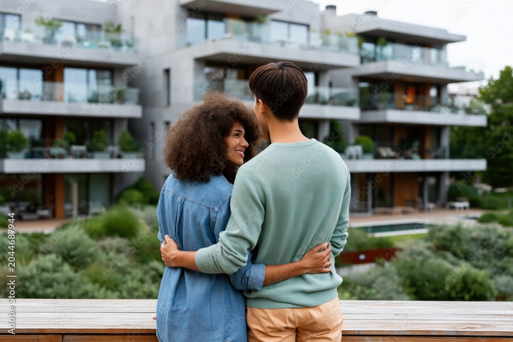 Fototapeta premium Smiling couple embracing on balcony with contemporary apartments in background for lifestyle banner and relationship design