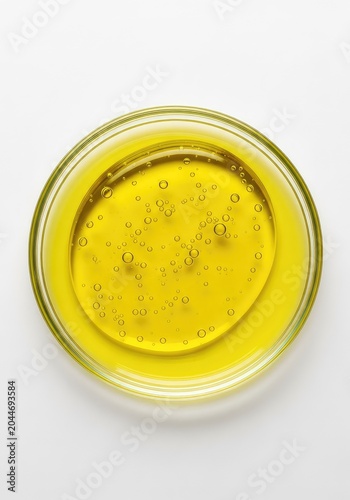 Macro view of a vibrant yellow oil swatch inside a glass petri dish with tiny oxygen bubbles for cosmetic science on a white background, spa, concentrated, glossy