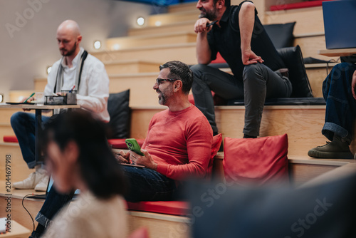 Fotografija A group of men attend a casual business meeting in tiered seating, some taking n