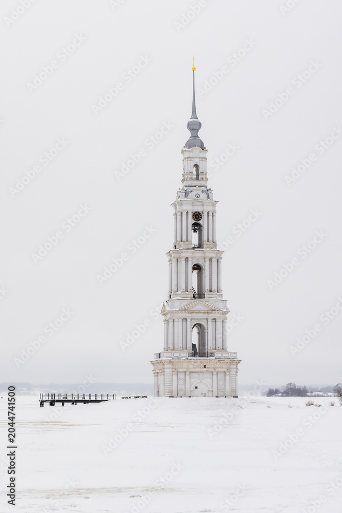 Obraz premium Vertical photo of a tall white bell tower rises alone on a frozen, snow-covered Volga river beneath an overcast winter sky. Landscape with the Flooded Belfry, Kalyazin, Tver Oblast, Russia