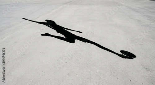 A dramatic long shadow stretches across a sunlit pavement, creating an abstract and elongated figure, symbolizing movement and the transient nature of light and form