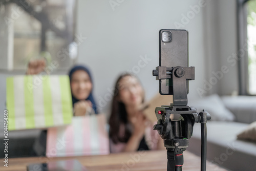 Smartphone mounted on a tripod films a casual review or unboxing scene with two blurred women holding shopping bags in a bright modern interior