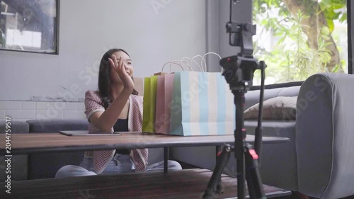 Asian woman unboxing colorful shopping bags behind camera, cozy living room, tripod smartphone, presenting products with smile,