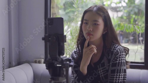 Asian woman creator filming product demo, adjusting phone on tripod, demonstrating features, pointing gestures, window light,