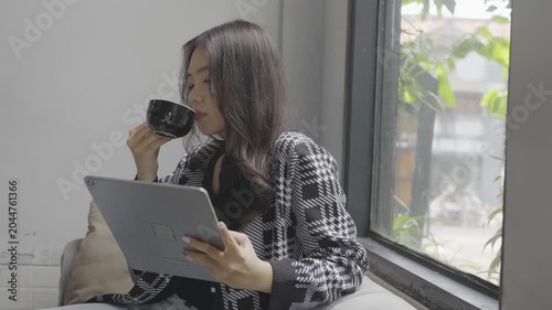 Asian woman reading tablet while sipping coffee by window, cozy sofa setup, plaid jacket, natural morning light, focused study