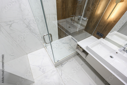 A modern bathroom with marble-patterned tiles, frameless glass shower enclosure, dual vessel sinks, wood-paneled accent wall, and chrome fixtures