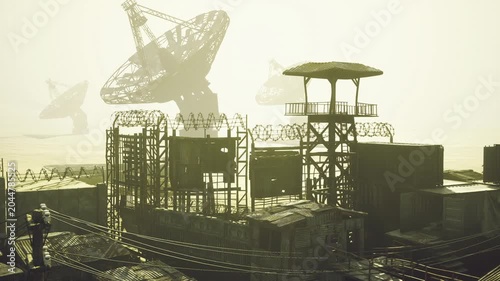 Coastal satellite relay at sunrise, stacked containers and watchtower silhouette, faint sea visible beyond, warm glare and atmospheric diffusion, quiet