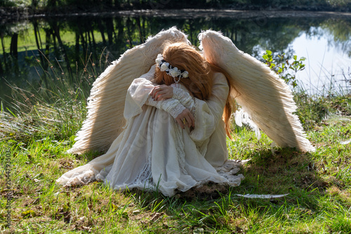 A female angel sits sadly in the fetal position on the shore of a small lake
