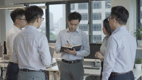 Group of Asian business professionals engaged in discussion, with one man taking notes in a notebook while others listen attentively in a modern office setting