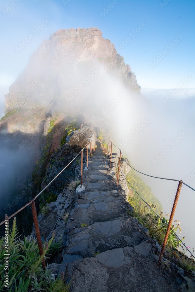 Fototapeta premium Narrow mountain trail between Pico do Arieiro and Pico Ruivo on Madeira Island, Portugal. Scenic hiking path overlooking a deep valley filled with inversion clouds and mist.