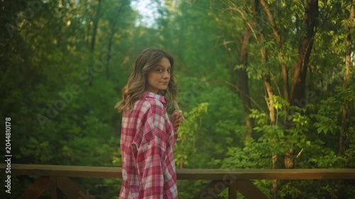 A young woman in a pink plaid shirt stands beside a wooden bridge railing in a lush forest, glancing back toward the camera with a calm expression. Soft