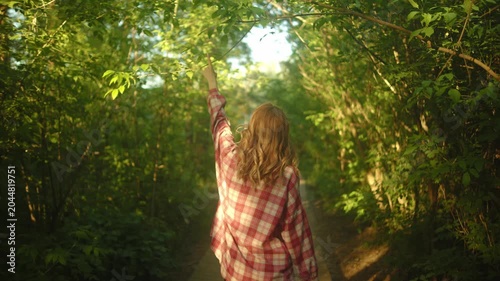 Rear view of a woman walking slowly on a narrow woodland trail surrounded by dense green foliage, soft sunlight, and a calm natural setting