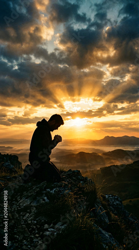 Silhouette of kneeling man in prayer on mountain top at sunset
