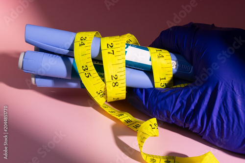Detailed view of a hand with blue latex glove holding semaglutide glp1 pens and a yellow tape measure on a bright pink background area.