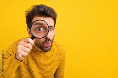 Young man examining through magnifying glass on yellow backdrop