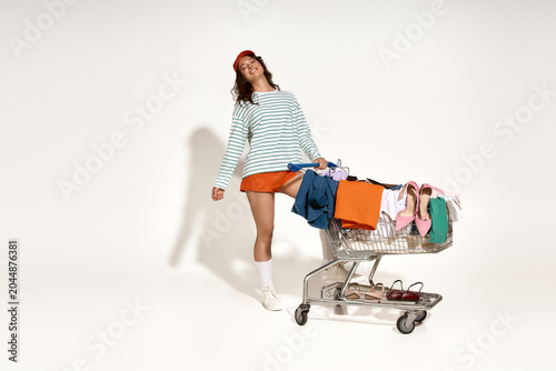 Tableau sur toile Stylish young woman joyfully shopping with a cart full of colorful clothes and a