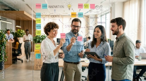A group of four business professionals discussing a project on a glass wall in an office setting.