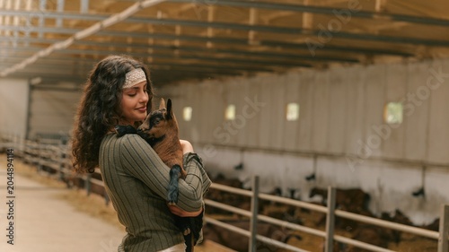 Canvastavla Young woman tenderly holding a small goat in a barn with other animals