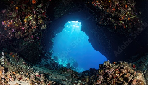 Sunbeams pierce a dark underwater cave, illuminating vibrant corals and clear blue depths