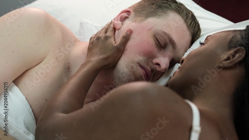 Sensual multiethnic couple kissing and hugging in bed at home. Romantic guy and girl having a good time together in the bedroom. A beautiful wife and a handsome husband look at each other, enjoy a