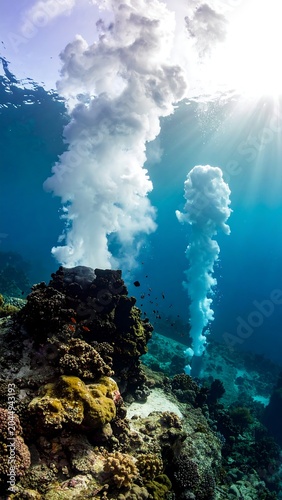 A vibrant underwater scene with sunlight shining through, illuminating two plumes of smoke or gas rising from a rocky, coral-covered seafloor