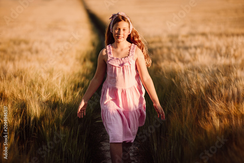 Fotografie Carefree Young Girl Walking Through Tall Golden Wheat Field in Pink Dress