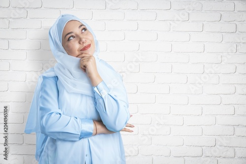 Muslim woman in blue hijab thinking and looking up against a white brick wall background